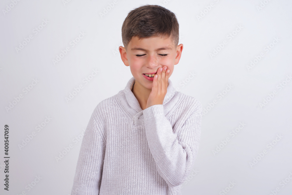 Cute Caucasian kid boy wearing knitted sweater against white wall touching mouth with hand with painful expression because of toothache or dental illness on teeth. Cute Caucasian kid boy wearing knitted sweater against white wall touching mouth with hand with painful expression because of toothache or dental illness on teeth.