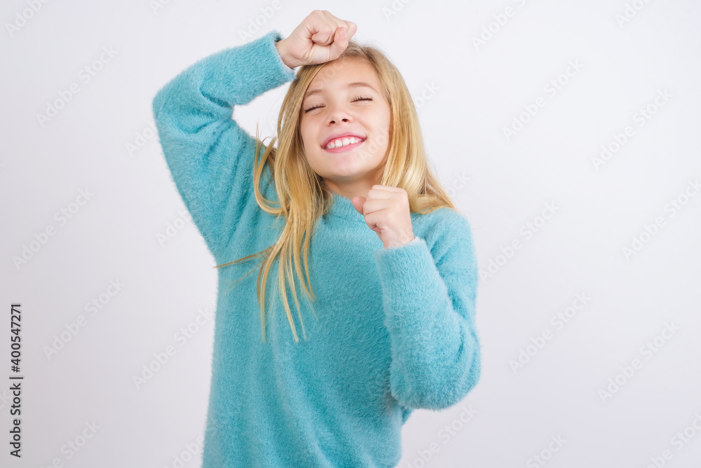 Attractive Cute Caucasian kid girl wearing blue knitted sweater against white wall celebrating a victory punching the air with his fists and a beaming toothy smile. Attractive Cute Caucasian kid girl wearing blue knitted sweater against white wall celebrating a victory punching the air with his fists and a beaming toothy smile.