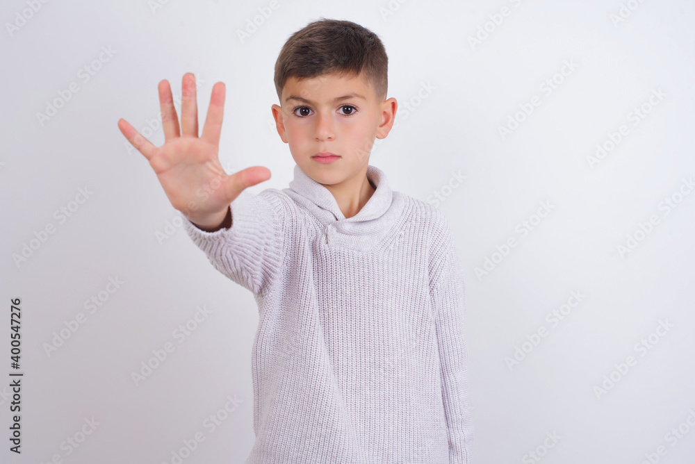 Cute Caucasian kid boy wearing knitted sweater against white wall doing stop sing with palm of the hand. Warning expression with negative and serious gesture on the face. Cute Caucasian kid boy wearing knitted sweater against white wall doing stop sing with palm of the hand. Warning expression with negative and serious gesture on the face.