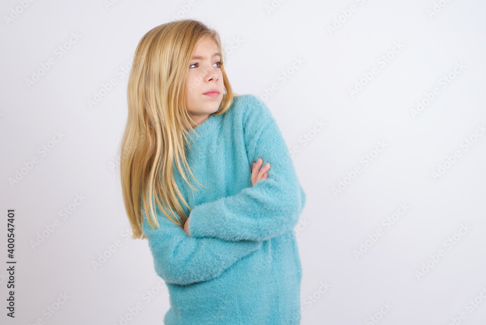 Image of upset Cute Caucasian kid girl wearing blue knitted sweater against white wall with arms crossed. Looking with disappointed expression aside after listening to bad news. Image of upset Cute Caucasian kid girl wearing blue knitted sweater against white wall with arms crossed. Looking with disappointed expression aside after listening to bad news.