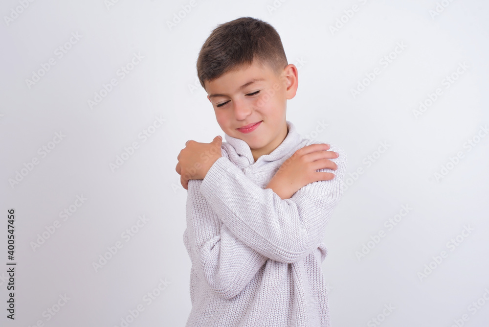Cute Caucasian kid boy wearing knitted sweater against white wall Hugging oneself happy and positive, smiling confident. Self love and self care Cute Caucasian kid boy wearing knitted sweater against white wall Hugging oneself happy and positive, smiling confident. Self love and self care