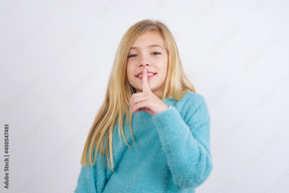 Smiling Cute Caucasian kid girl wearing blue knitted sweater against white wall makes shush gesture, holds fore finger over lips hides secret. Be mute, please. Smiling Cute Caucasian kid girl wearing blue knitted sweater against white wall makes shush gesture, holds fore finger over lips hides secret. Be mute, please.