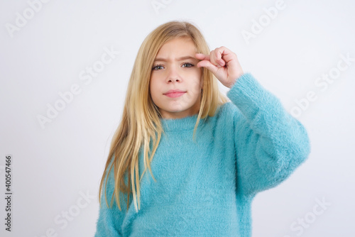 Upset Cute Caucasian kid girl wearing blue knitted sweater against white wall shapes little gesture with hand demonstrates something very tiny small size. Not very much © Jihan