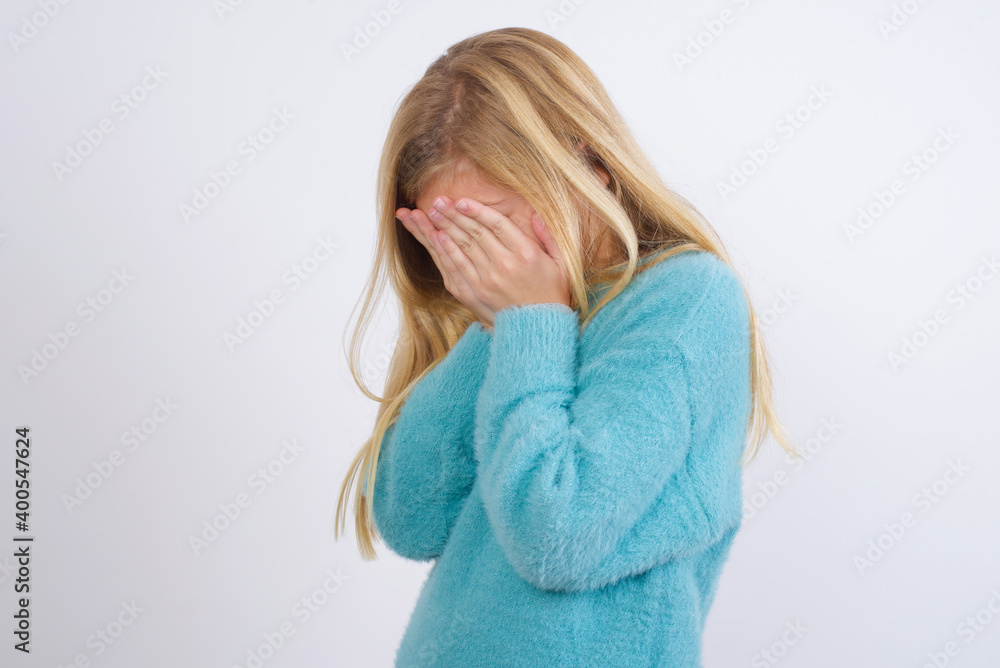 Sad Cute Caucasian kid girl wearing blue knitted sweater against white wall covering face with hands and crying. Sad Cute Caucasian kid girl wearing blue knitted sweater against white wall covering face with hands and crying.