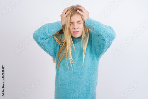 Cute Caucasian kid girl wearing blue knitted sweater against white wall holding head with hands, suffering from severe headache, pressing fingers to temples