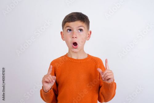 Fotografia Cute Caucasian kid boy wearing knitted sweater against white wall amazed and surprised looking up and pointing with fingers and raised arms