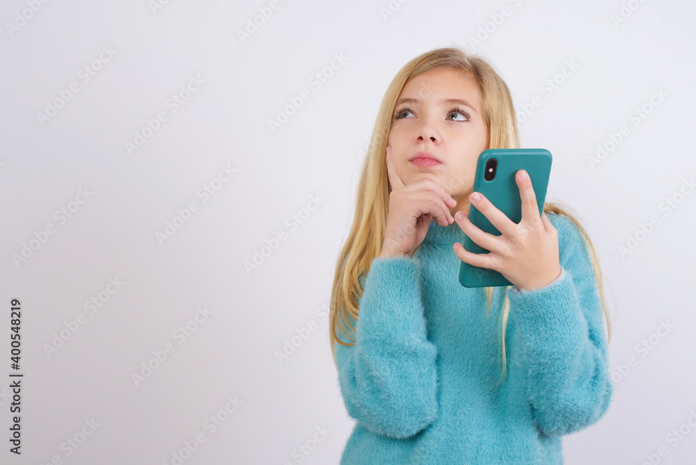 Thinking dreaming Cute Caucasian kid girl wearing blue knitted sweater against white wall using mobile phone and holding hand on face. Taking decisions and social media concept. Thinking dreaming Cute Caucasian kid girl wearing blue knitted sweater against white wall using mobile phone and holding hand on face. Taking decisions and social media concept.