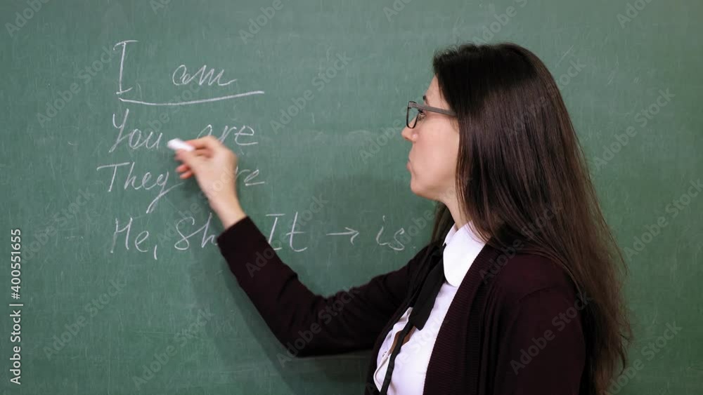Online schooling. Female teacher is holding online English lesson. She is writing grammar rules with chalk on blackboard, in school class. remote teaching. live video streaming from class. Online schooling. Female teacher is holding online English lesson. She is writing grammar rules with chalk on blackboard, in school class. remote teaching. live video streaming from class.