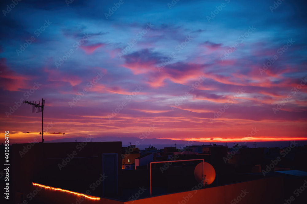 Sun appears from behind the clouds, above the city, roof top view. Summer sunrise or sunset. Dark blue and red sky color tones. Old town of Spain.