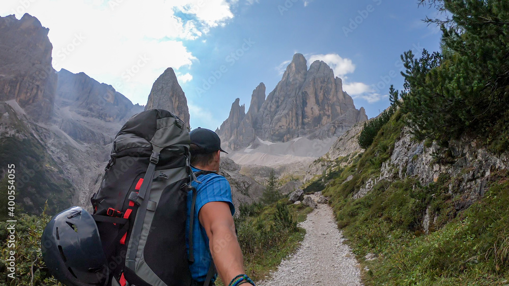 A man with big backpack taking a selfie while hiking along a narrow pathway in Italian Dolomites. There is a massive mountain with very steep and sharp slopes. Smaller mountains around. Raw landscape