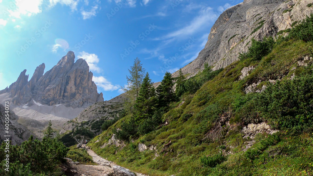 A sunny and warm day in Italian Dolomites. The slopes are overgrown with lush green plants. Few taller trees along the pathway. In the back, there are sharp and high mountain chains. Experiencing