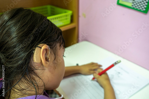 A girl wearing a hearing aid and sitting and writing a book in the classroom.