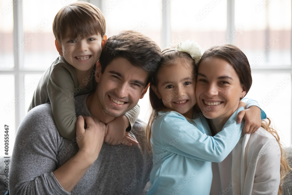 Head shot portrait smiling parents with two adorable little kids, happy father piggy backing preschool son, cute girl hugging beautiful young mother, family posing for photo at home, enjoying weekend Head shot portrait smiling parents with two adorable little kids, happy father piggy backing preschool son, cute girl hugging beautiful young mother, family posing for photo at home, enjoying weekend