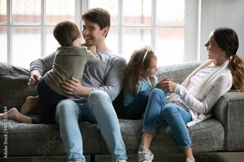 Happy parents with two children having fun at home, tickling, sitting on cozy couch at home, overjoyed mother and father cuddling with adorable little daughter and son, enjoying funny activity