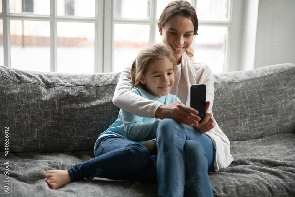 Close up happy mother and little daughter having fun with phone together, sitting on cozy couch at home, smiling mum hugging adorable child cute girl, making video call to relatives, browsing apps Close up happy mother and little daughter having fun with phone together, sitting on cozy couch at home, smiling mum hugging adorable child cute girl, making video call to relatives, browsing apps