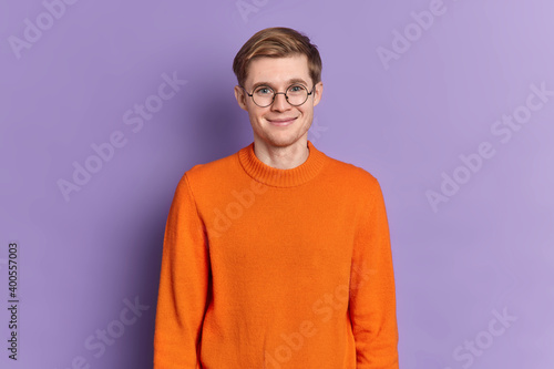 Portrait of handsome European male student has gentle smile on face happy to hear pleasant news stands delighted wears round spectacles orange jumper isolated over purple background. Happy youngster