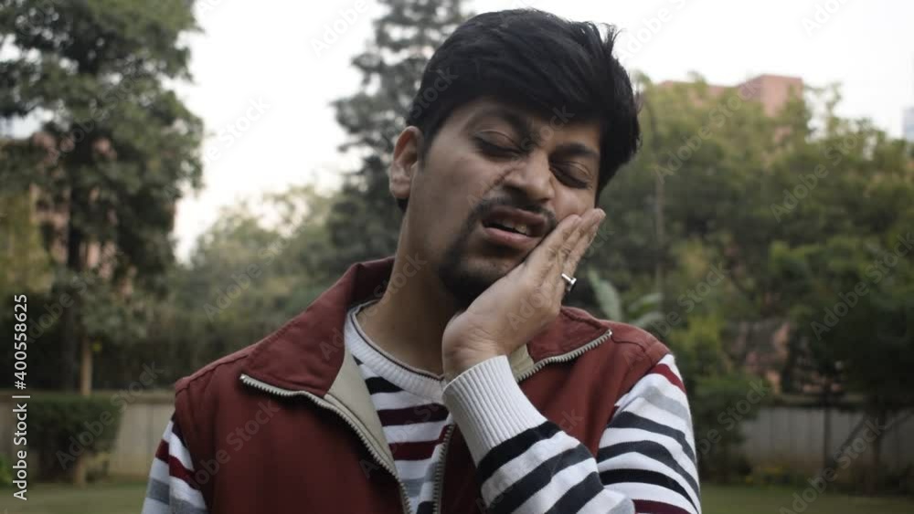 Young Indian boy making faces due to tooth ache and oral problems. He is wearing stripped Shirt and is standing in a park Young Indian boy making faces due to tooth ache and oral problems. He is wearing stripped Shirt and is standing in a park