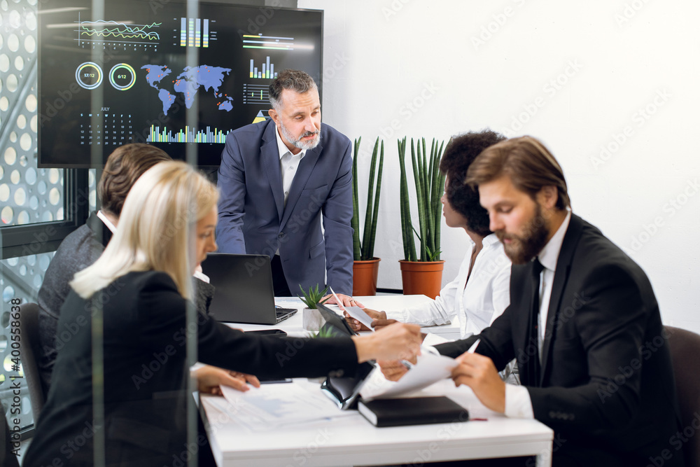 Diverse multiethnic team of executive directors with mature male boss, on a meeting in conference room, sharing opinions, working with papers, tablets and statistics charts on huge TV screen behind Diverse multiethnic team of executive directors with mature male boss, on a meeting in conference room, sharing opinions, working with papers, tablets and statistics charts on huge TV screen behind