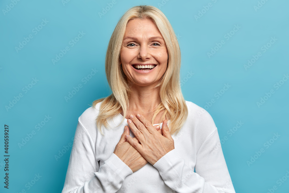 Photo of tender blonde woman with wrinkles and blonde hair keeps hands pressed to chest being sincerely thankful for good supporitve words smiles gently wears casual white jumper isolated on blue Photo of tender blonde woman with wrinkles and blonde hair keeps hands pressed to chest being sincerely thankful for good supporitve words smiles gently wears casual white jumper isolated on blue