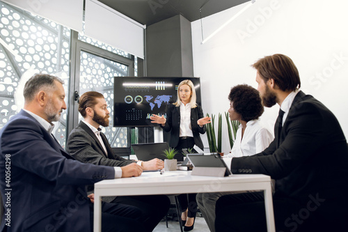 Attractive confident blond mature lady in formal suit, gesturing and reporting about ecological, economic or marketing problems to her multiethnic team of colleagues in boardroom during meeting
