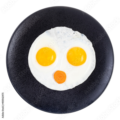 top view of fried eggs and slice of carrot on black plate isolated on white background. Fried eggs like face with open mouth
