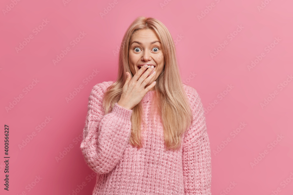 Portrait of blonde very happy surprised woman covers mouth and looks with unbelievable gaze at camera wears knitted sweater poses against rosy background. People and human reactions concept. Portrait of blonde very happy surprised woman covers mouth and looks with unbelievable gaze at camera wears knitted sweater poses against rosy background. People and human reactions concept.