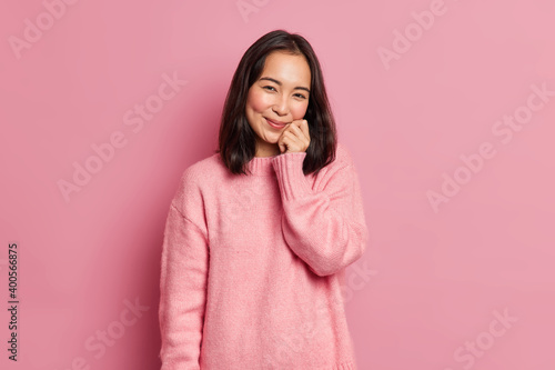 Bild auf Leinwand Lovely brunette woman with eastern appearance keeps hand on face has gentle smile looks satisfied at camera has dimples on rouge cheeks poses against pink background