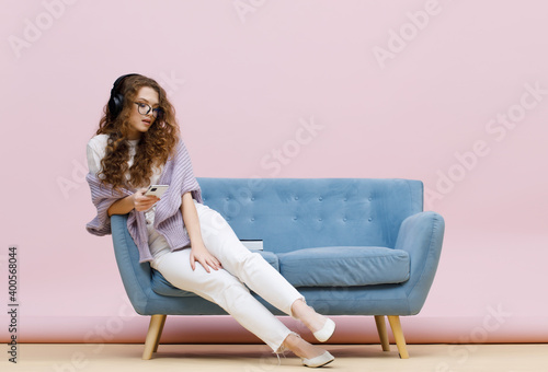 Beautiful curly-haired girl in casual clothes and glasses sits on sofa. Pastel trendy pink background. Portrait of smiling young woman in headphones listening music with smartphone in hands.
