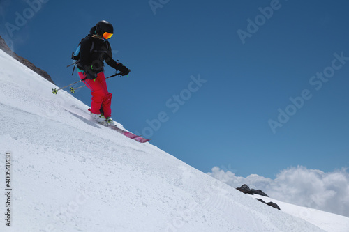 woman skier athlete makes a jump in flight on a snowy slope against the backdrop of a blue sky of mountains and clouds. Freeride and extreme skiing for women