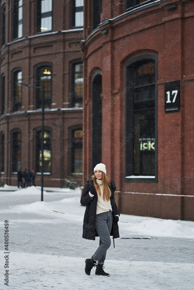 a young woman stands in a light warm outerwear in winter near a brick loft building with tall black windows