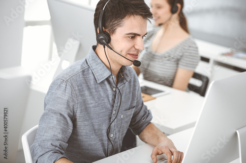 Casual dressed young man using headset and computer while talking with customers online. Group of operators at work. Call center