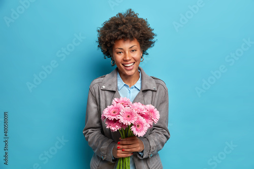 Studio shot of happy culy haired woman holds beautiful flowers receives her favorite gerberas from beloved person wears fashionable grey jacket isolated over blue background. Feminine concept