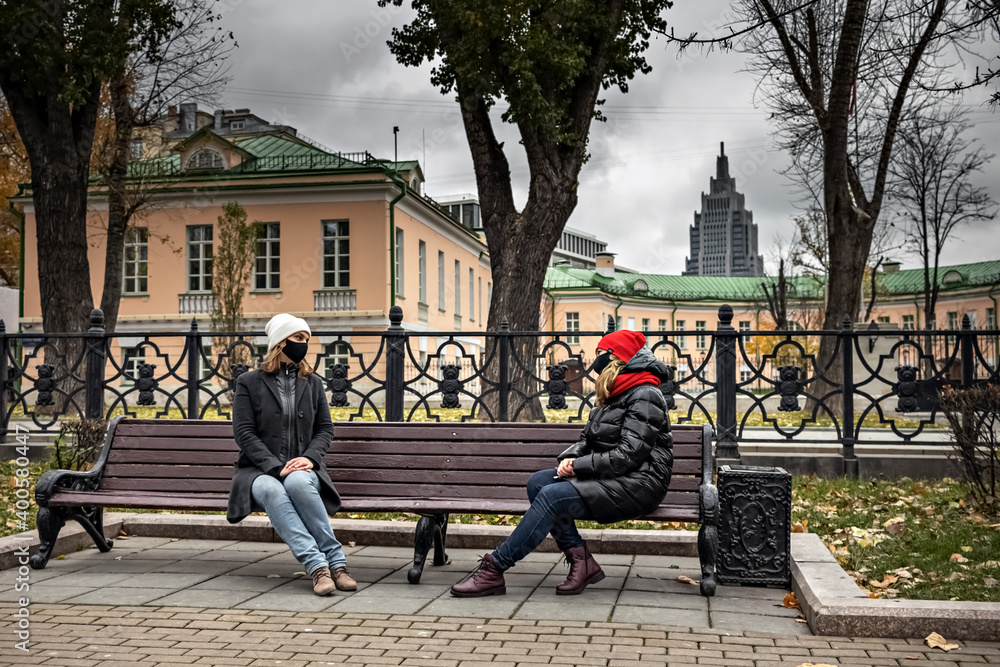 Two young women wearing medical masks talking at a safe social distance while sitting on a bench in an autumn park. Meeting and chatting with friends during the coronavirus pandemic