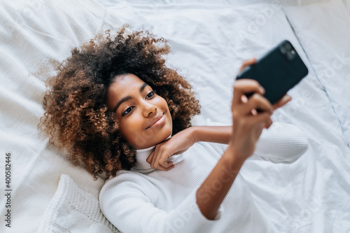 Lady with short kinky hair and dark skin lying on bed and filming beauty blog on smartphone in modern bedroom closeup