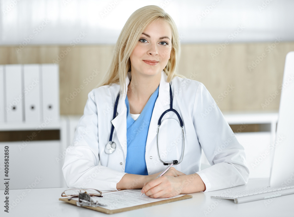 Woman-doctor at work while sitting at the desk in hospital or clinic. Blonde cheerful physician filling up medication history record form Woman-doctor at work while sitting at the desk in hospital or clinic. Blonde cheerful physician filling up medication history record form