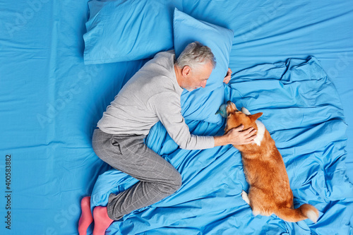 Photo of happy aged bearded man in nightwear plays with dog expresses love and care pose together on comfortable bed have friendly relationships Male pensioner enjoys lazy day with pet at home