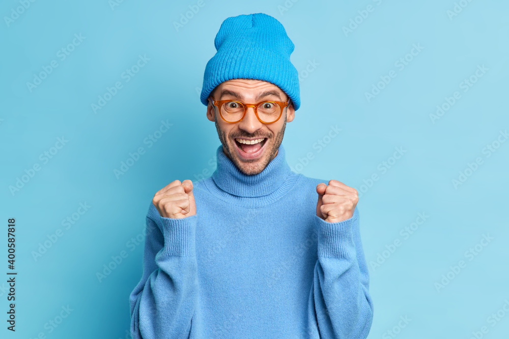 Overjoyed unshaven European guy raises clenched fists rejoices triumph feels like winner feels joy wears casual turtleneck and hat isolated over blue background. People happiness success concept Overjoyed unshaven European guy raises clenched fists rejoices triumph feels like winner feels joy wears casual turtleneck and hat isolated over blue background. People happiness success concept