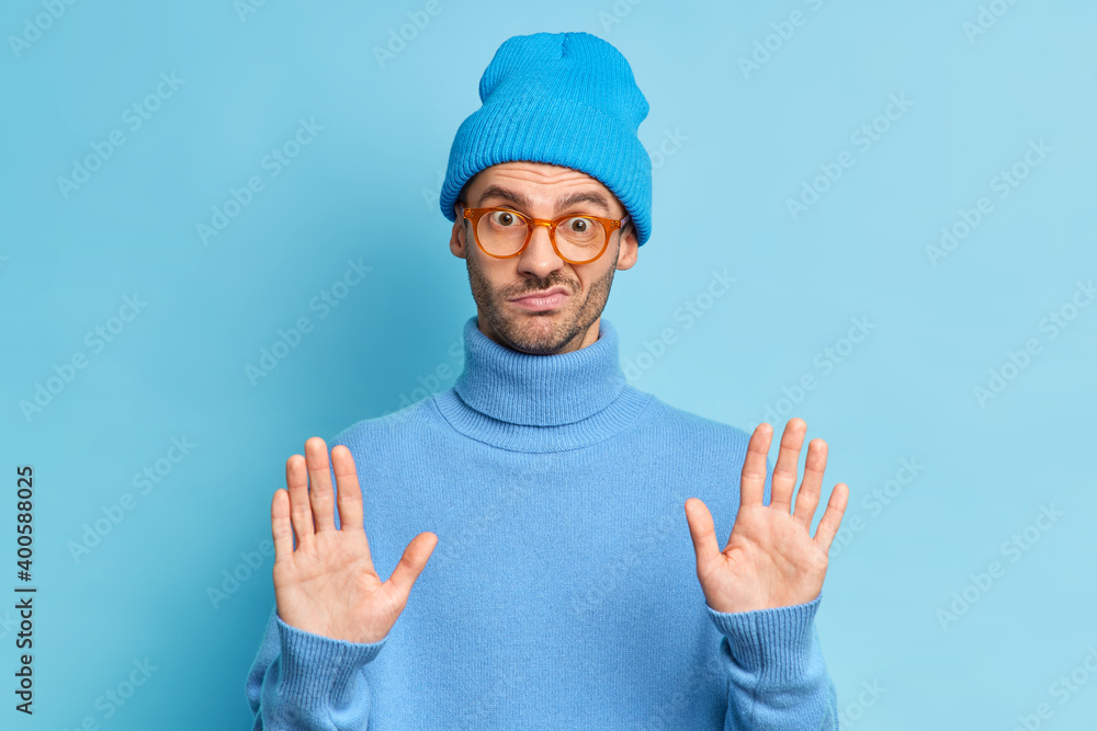 Serious handsome man with bristle keeps palms forward camera refuses something or makes decline gesture wears blue hat and turtleneck poses indoor. Oh no I dont agree on this. Stop right now Serious handsome man with bristle keeps palms forward camera refuses something or makes decline gesture wears blue hat and turtleneck poses indoor. Oh no I dont agree on this. Stop right now