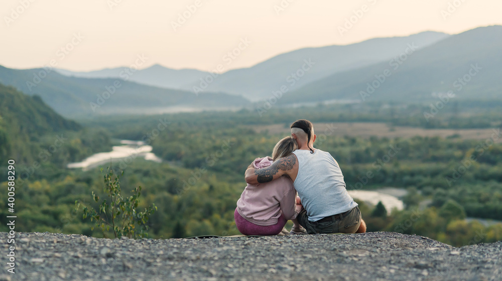 Young man hugs his girlfriend on the mountain peak at sunset background.