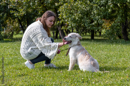 Beautiful caucasian woman in white sweater strokes labrador puppy during dog walking in city park on a sunny day. The puppy is about 5 months old. Pets theme.