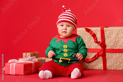 Cute little baby in elf's costume and with gifts on color background