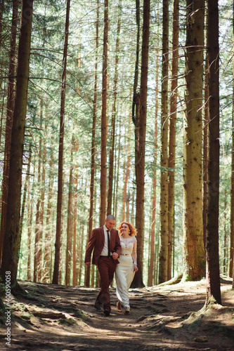 Happy stylish couple newlyweds in the green forest on summer day. bride in long white dress and groom in red suit are hugging. wedding day. © Andriy Medvediuk