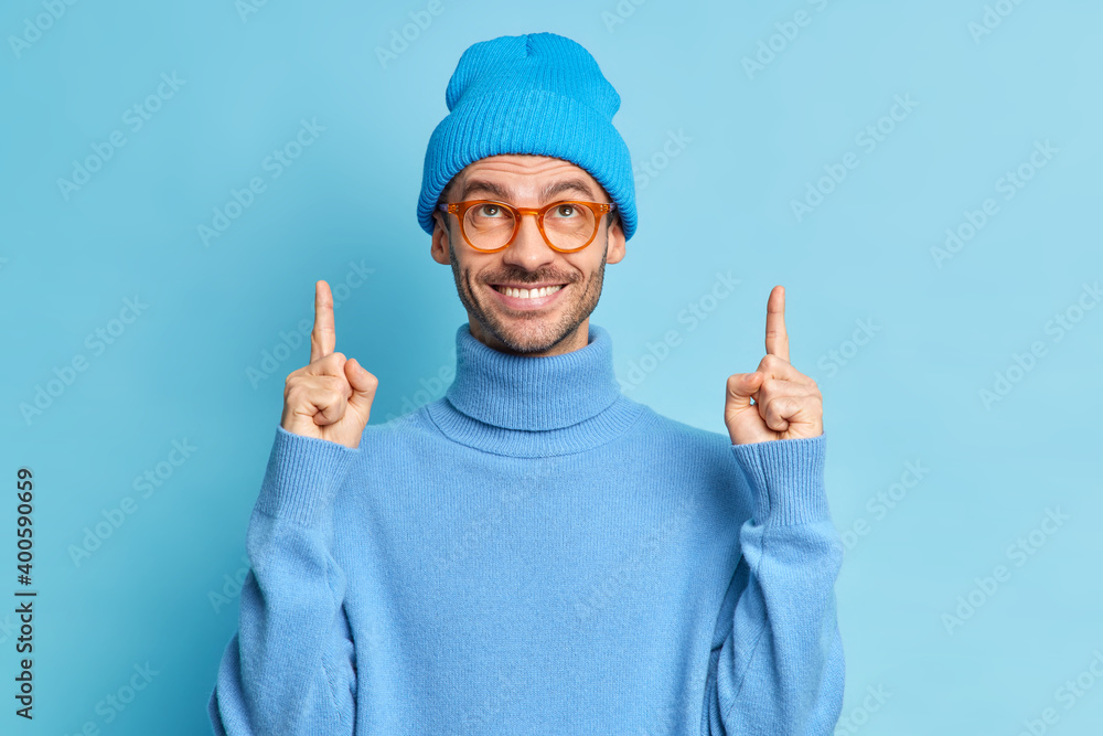 Positive handsome male youngster with bristle smiles and indicates with both index fingers upwards demonstrates nice offer for you wears blue hat and turtleneck poses indoor. Check this out.