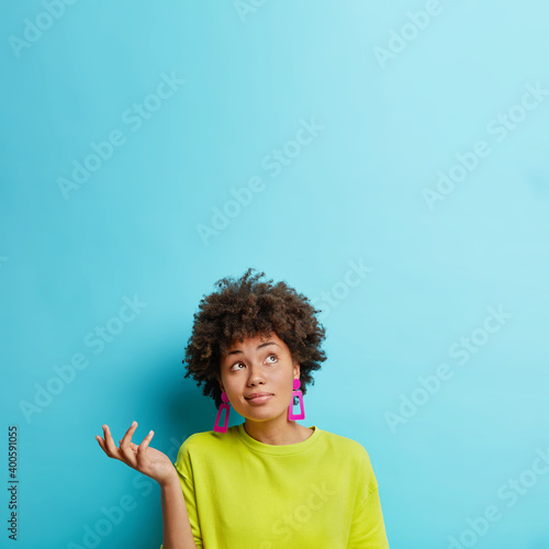 Vertical shot of doubtful Afro American woman raises hand with doubt looks confused above makes decision dressed casually isolated over blue background blank space for your promotional content photo