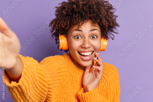 Lovely cheerful dark skinned woman smiles broadly has fun indoor makes selfie dressed in casual orange sweater listens favorite music poses happy against purple background. People lifestyle concept