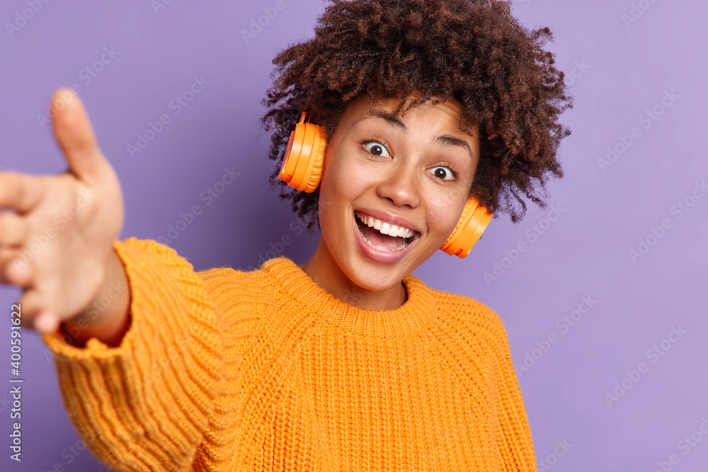 Positive dark skinned ethnic woman smiles broadly and stretches arm has fun indoor wears headphones on ears listens pleasant music dressed in knitted orange jumper poses against purple background Positive dark skinned ethnic woman smiles broadly and stretches arm has fun indoor wears headphones on ears listens pleasant music dressed in knitted orange jumper poses against purple background