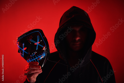 Man in hoodie with blurred face hold neon glowing scary mask. Anonymous concept