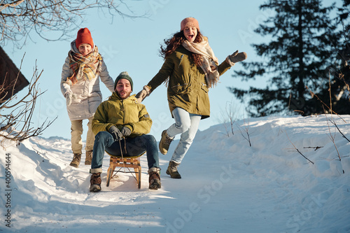 Fototapeta Joyful girls in winterwear running after young man on sledge moving down hill