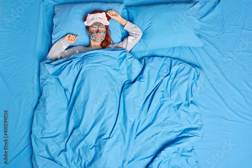 Photo of relaxed woman fell asleep while undergoing beauty procedures sleeps pacefully in bed with clay mask on face wears sleepmask and pajama poses on back being alone enjoys cozy atmosphere photo