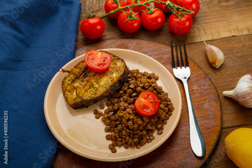 A plate of fish with lentils and tomatoes on a round stand with a fork on a blue napkin